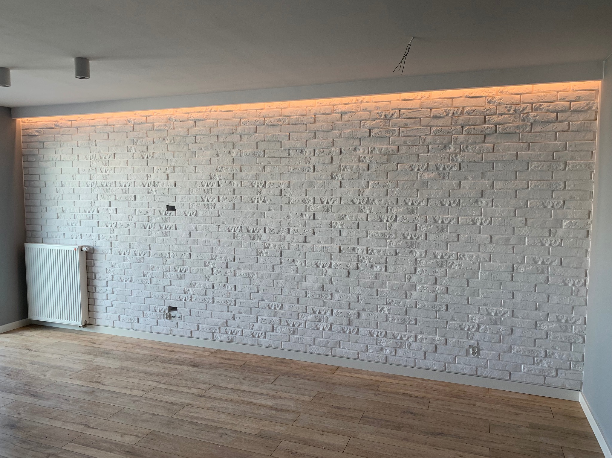 Biała ściana z cegły dekoracyjnej, oświetlona od góry ciepłym światłem LED, z widocznym grzejnikiem i drewnianą podłogą. Gniazdka elektryczne w ścianie.