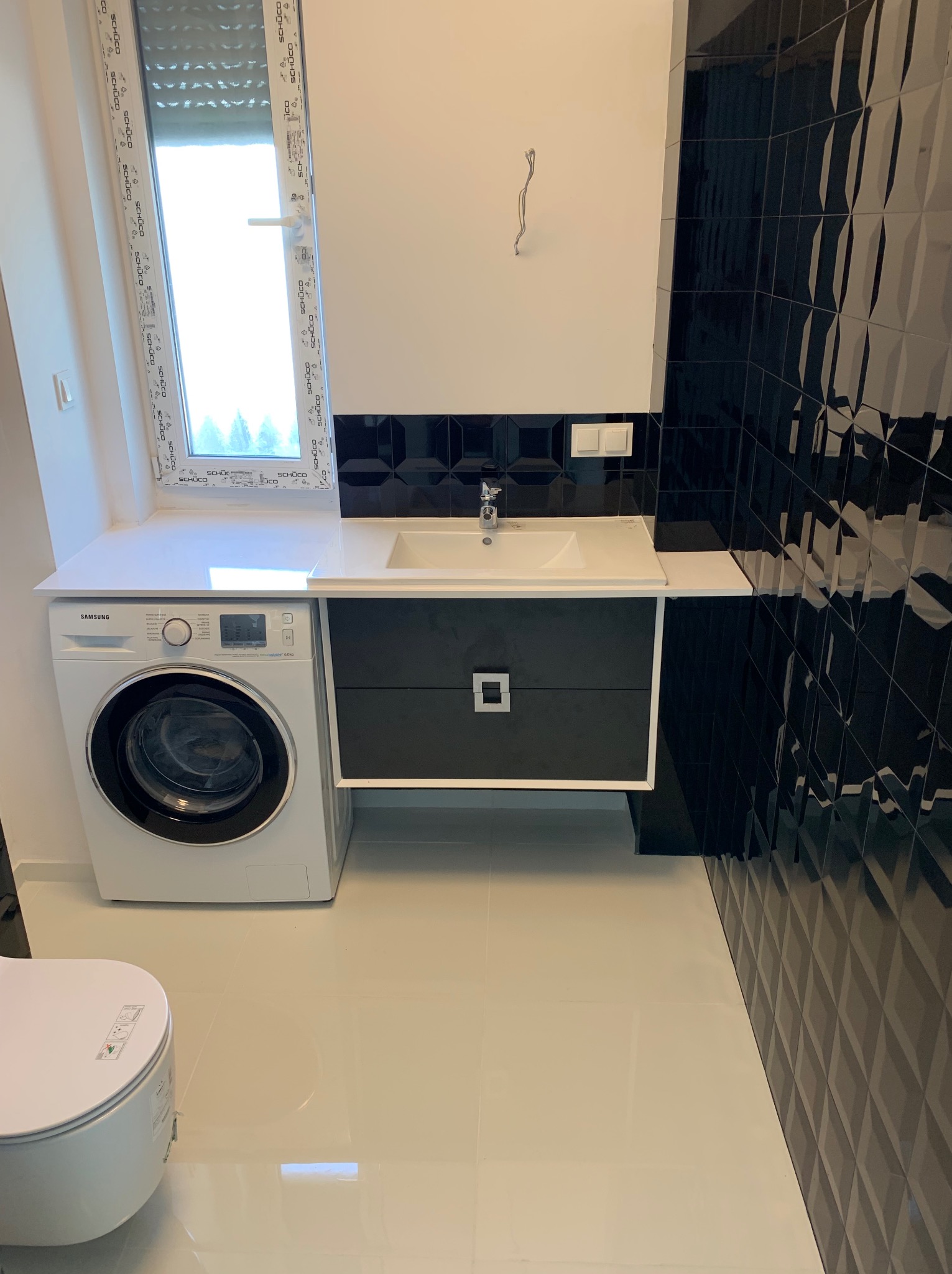 Nowoczesna łazienka po remoncie: białe płytki podłogowe, czarne, błyszczące płytki na ścianie, pralka Samsung obok umywalki z szafką. Okno z widokiem na zieleń.