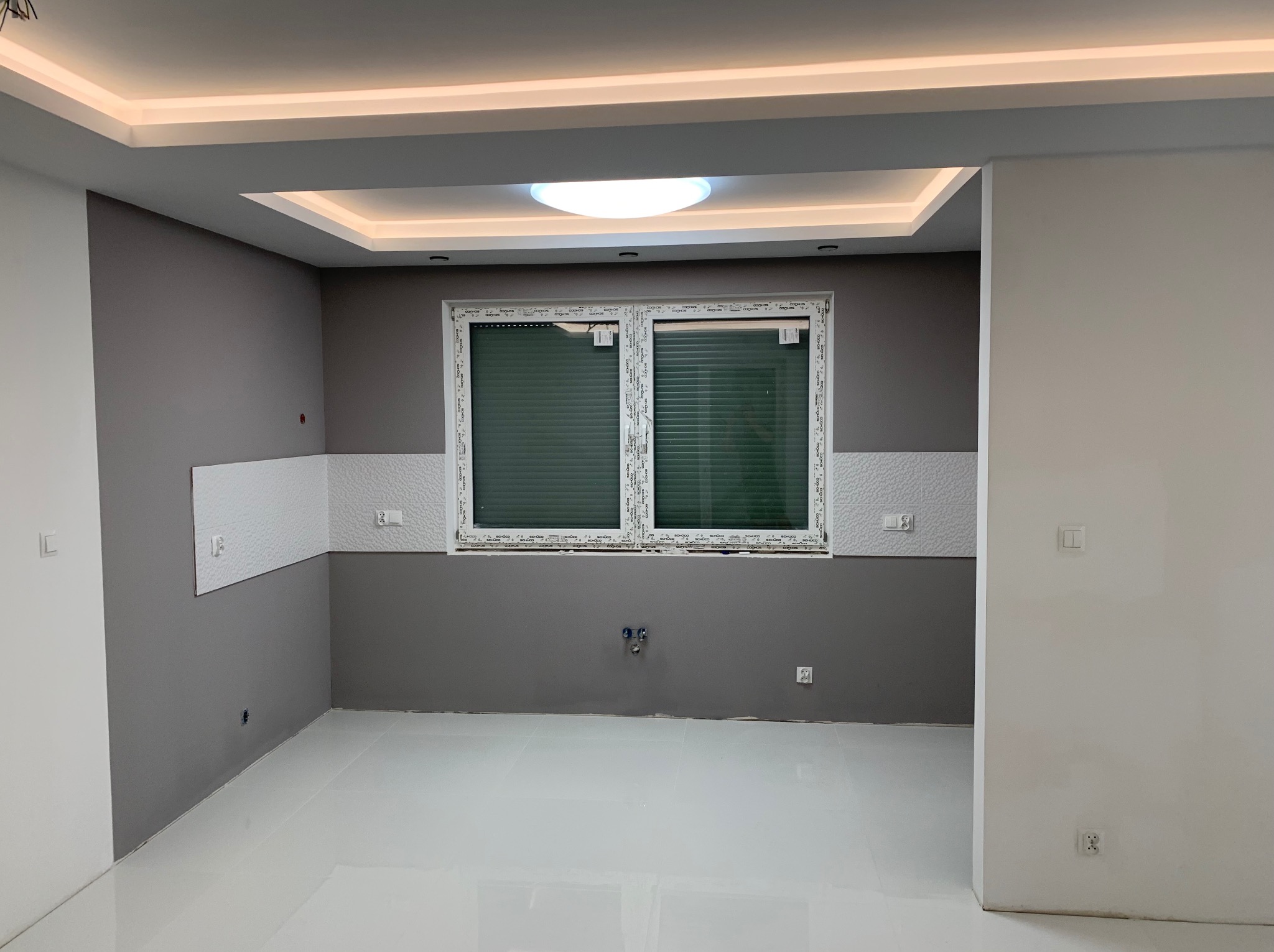 Wykończone wnętrze: szare ściany z białymi panelami, biała podłoga z połyskiem, okno z żaluzjami i nowoczesne oświetlenie sufitowe. Minimalistyczny design.