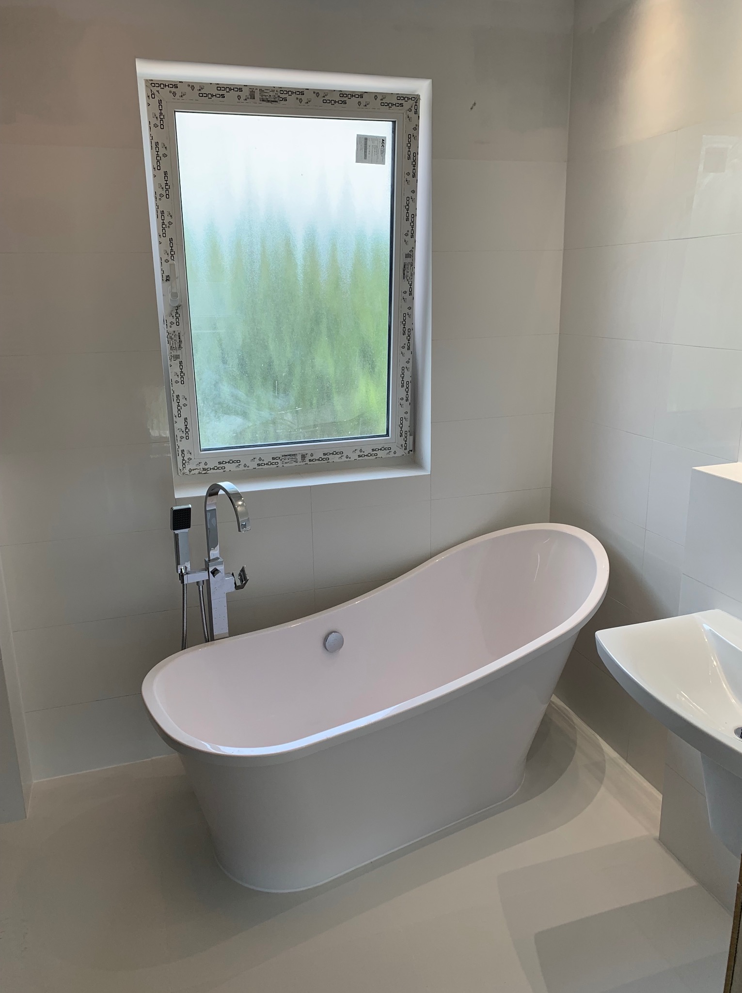 Nowoczesna, jasna łazienka po remoncie: wolnostojąca wanna, umywalka i okno z widokiem na zieleń. Białe płytki na ścianach i podłodze. Minimalistyczny styl.