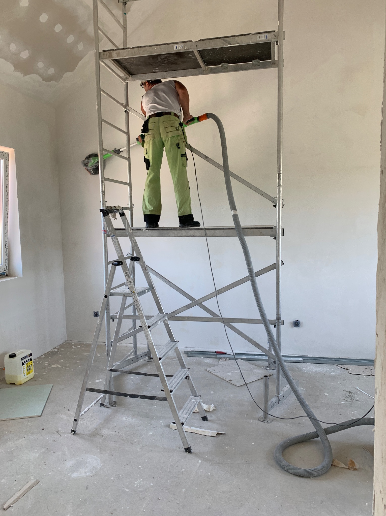 Pracownik na rusztowaniu szlifuje ścianę w jasnym wnętrzu. Widoczny odkurzacz przemysłowy i drabina. Ściany przygotowane do malowania. Etap prac wykończeniowych.