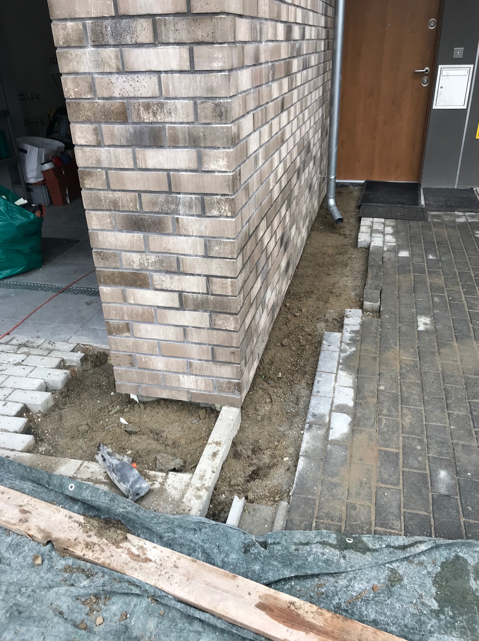 Układanie kostki brukowej przy ścianie budynku z cegły. Widoczne fragmenty ułożonej kostki, piasek i narzędzia. Prace brukarskie w toku, z bliska.