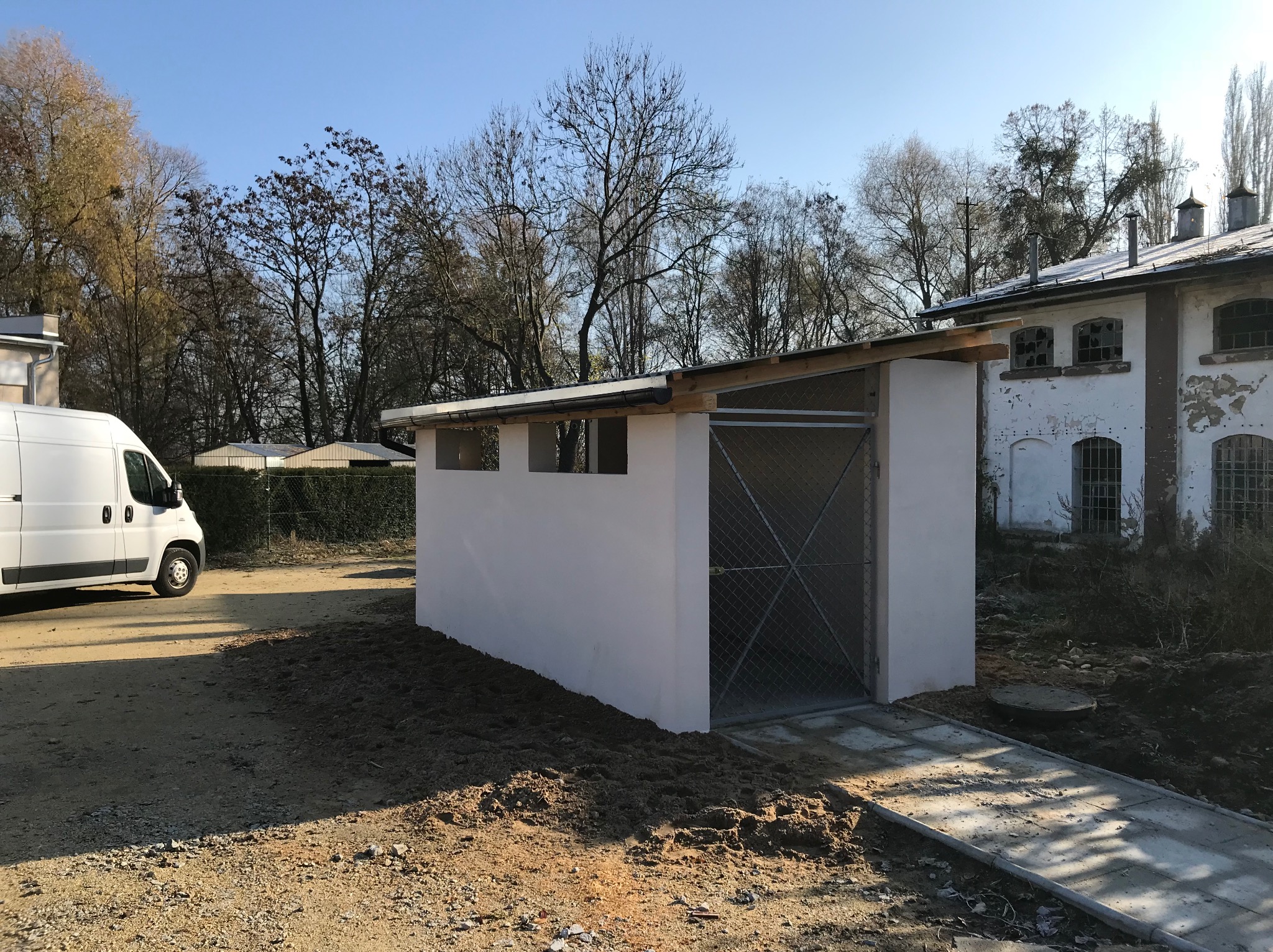 Nowo wybudowana, biała, betonowa konstrukcja z metalową bramą w tle budynku z cegły i białego vana. Surowy teren budowy z ziemią i gruzem.