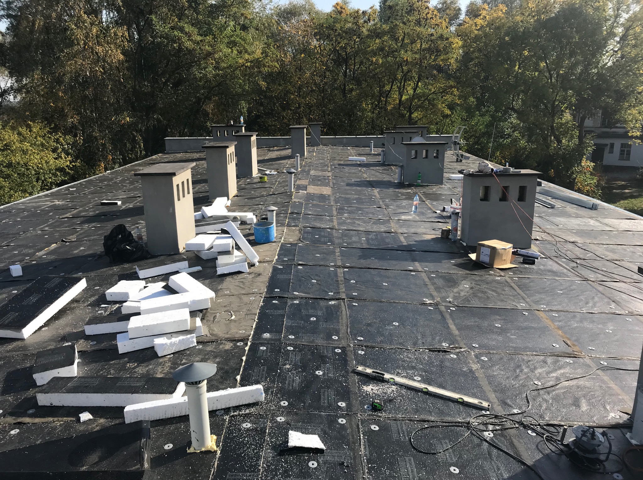 Czarny dach pokryty papą z kominami i fragmentami białego styropianu. Widoczne narzędzia i materiały budowlane. Prace dekarskie w toku, oświetlone naturalnym światłem.