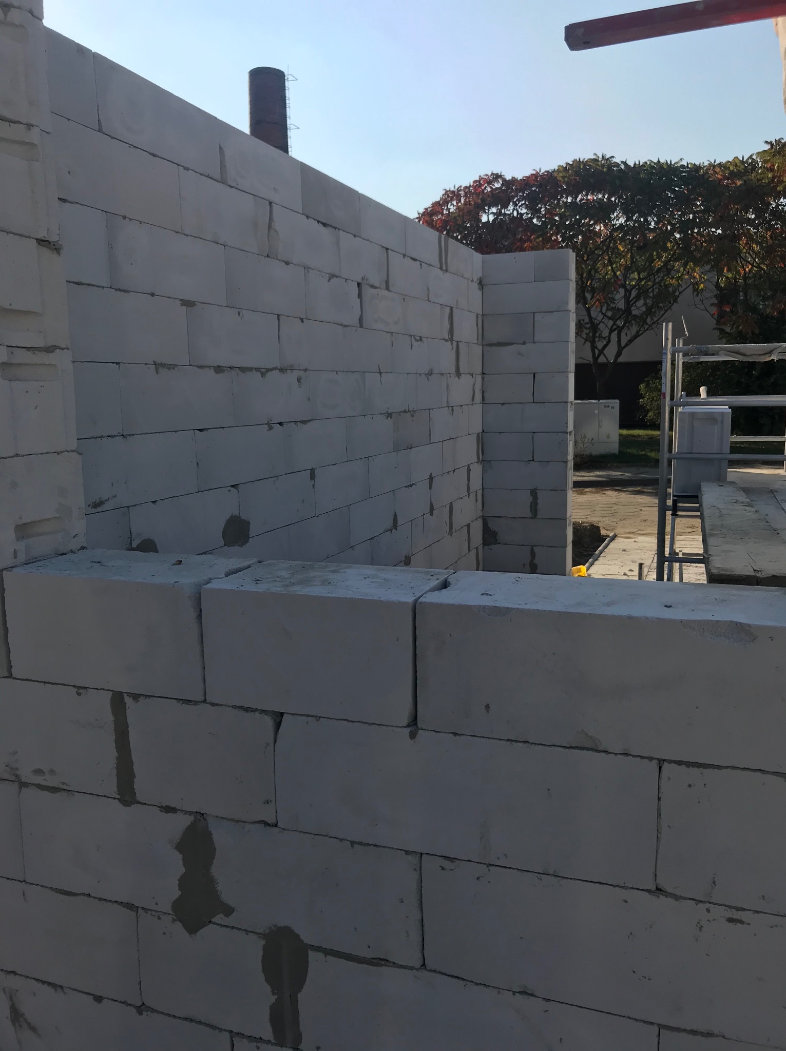 Ściana z białych bloczków, widoczne spoiny i fragment rusztowania. W tle zieleń drzew i błękitne niebo. Budowa domu w trakcie realizacji.