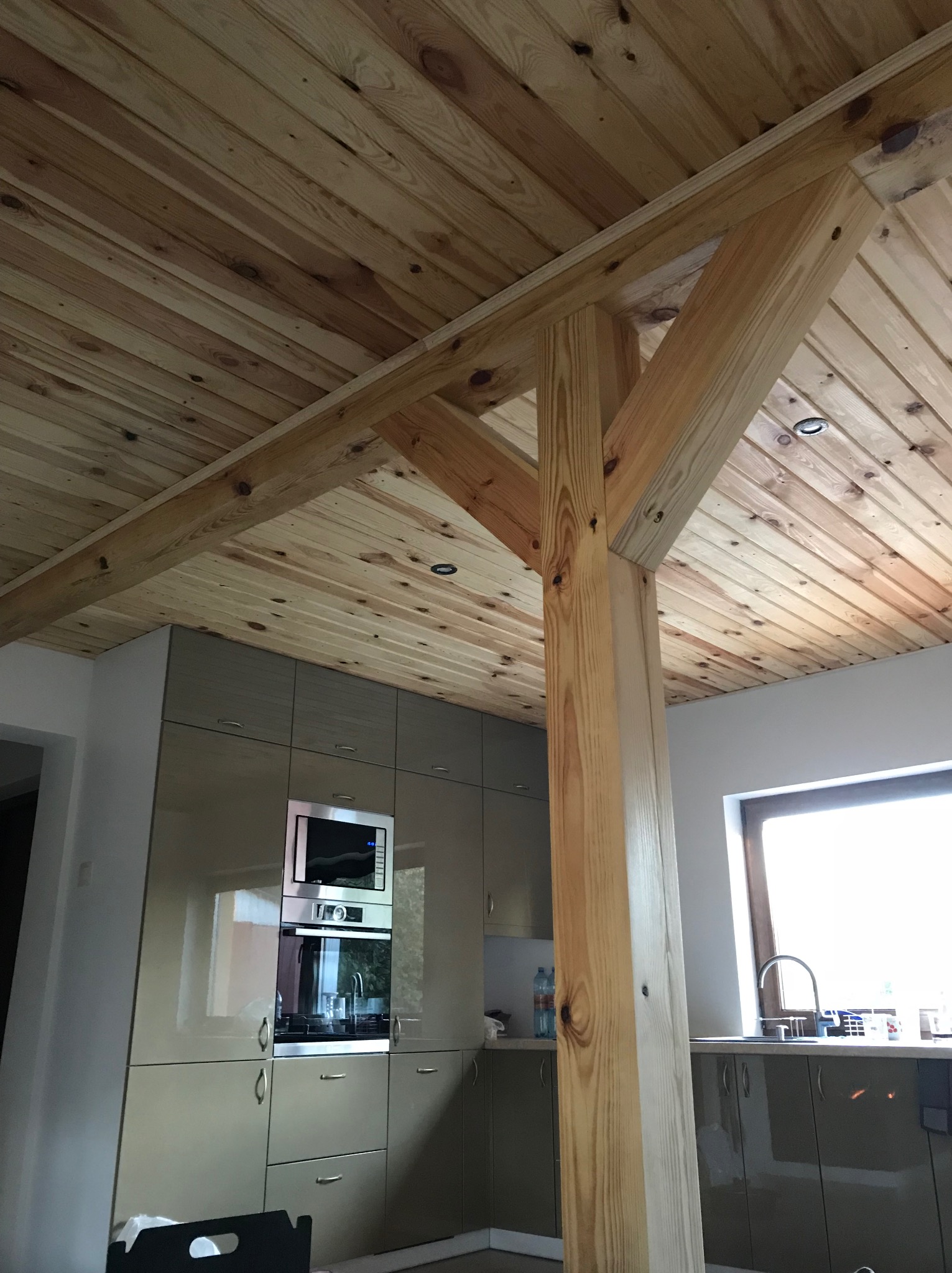 Drewniany sufit z belkami w jasnym odcieniu, widok z dołu na fragment kuchni z nowoczesnymi szafkami i oknem. Naturalne światło.