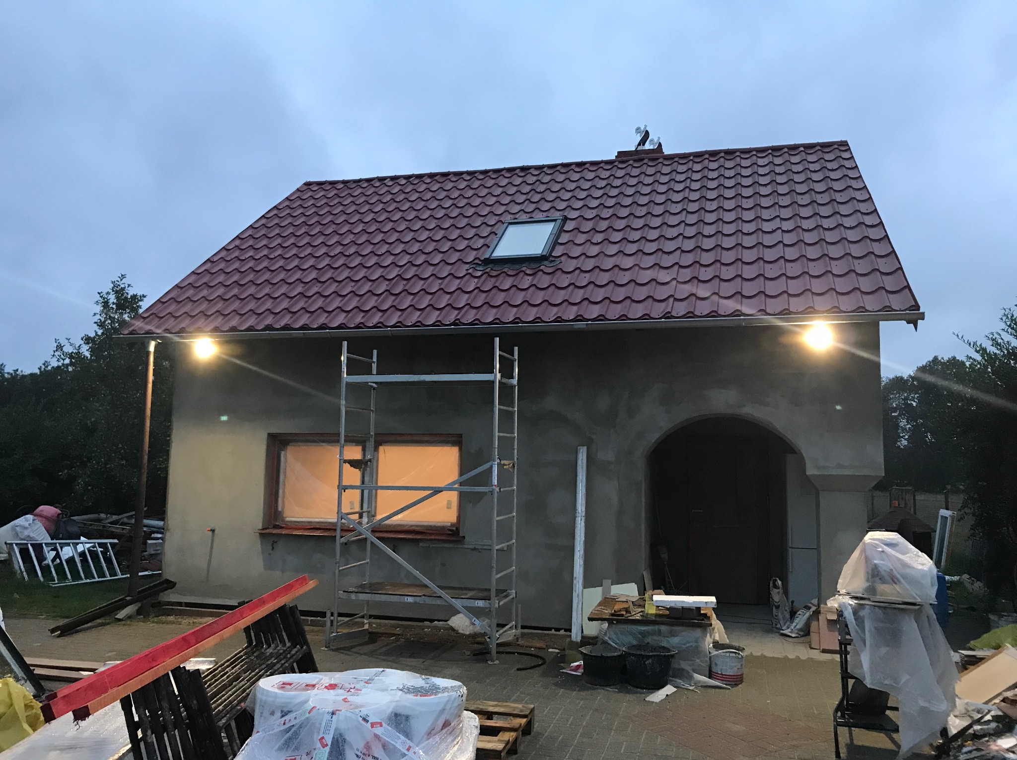 Dom w trakcie prac wykończeniowych: ocieplony budynek z nową, czerwoną dachówką, oknem dachowym i rusztowaniem. Widoczne materiały budowlane i oświetlenie zewnętrzne.