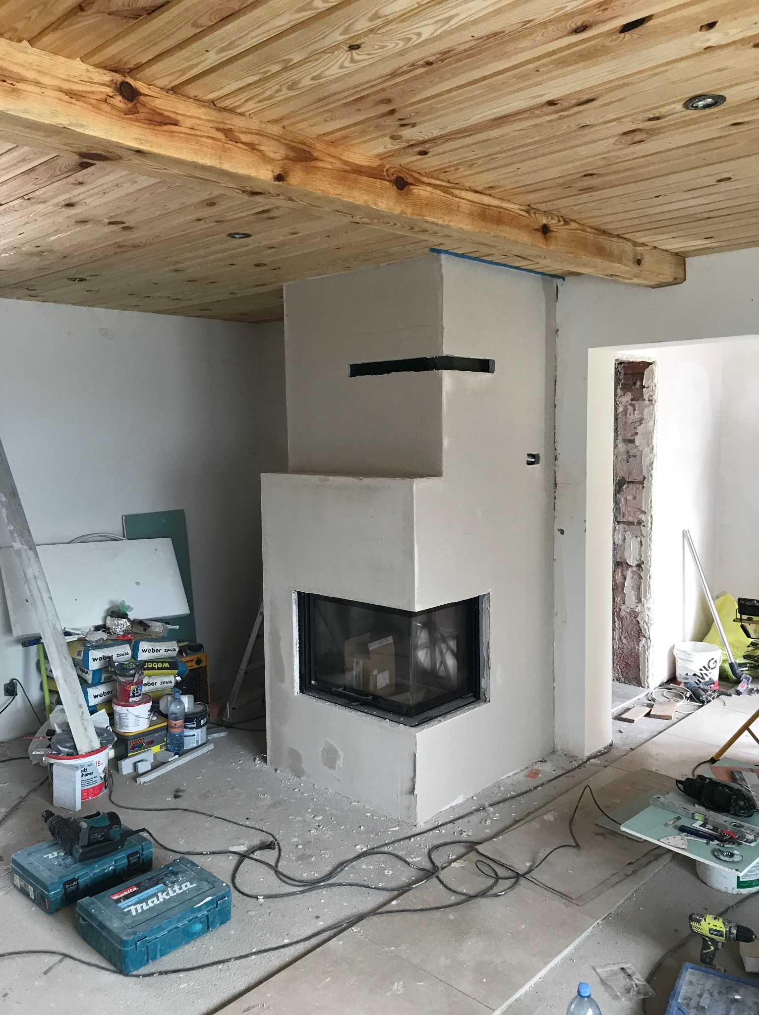 Wnętrze w trakcie wykończenia: obudowany kominek z wkładem, drewniany sufit, narzędzia i materiały budowlane. Widoczne puszki farb i narzędzia Makita.
