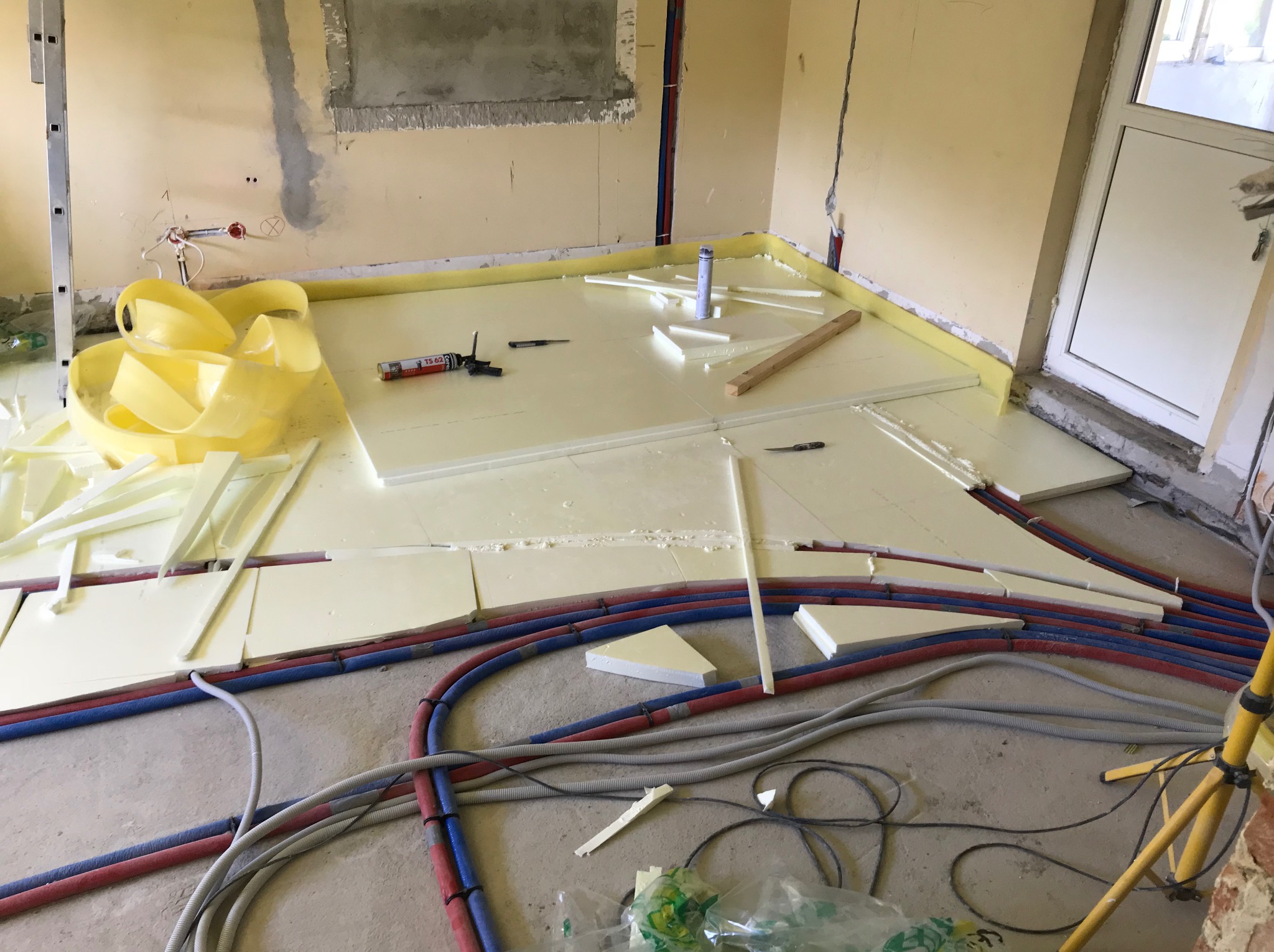 Układanie izolacji podłogowej z rurami grzewczymi w pomieszczeniu w trakcie remontu. Widoczne narzędzia i materiały izolacyjne.