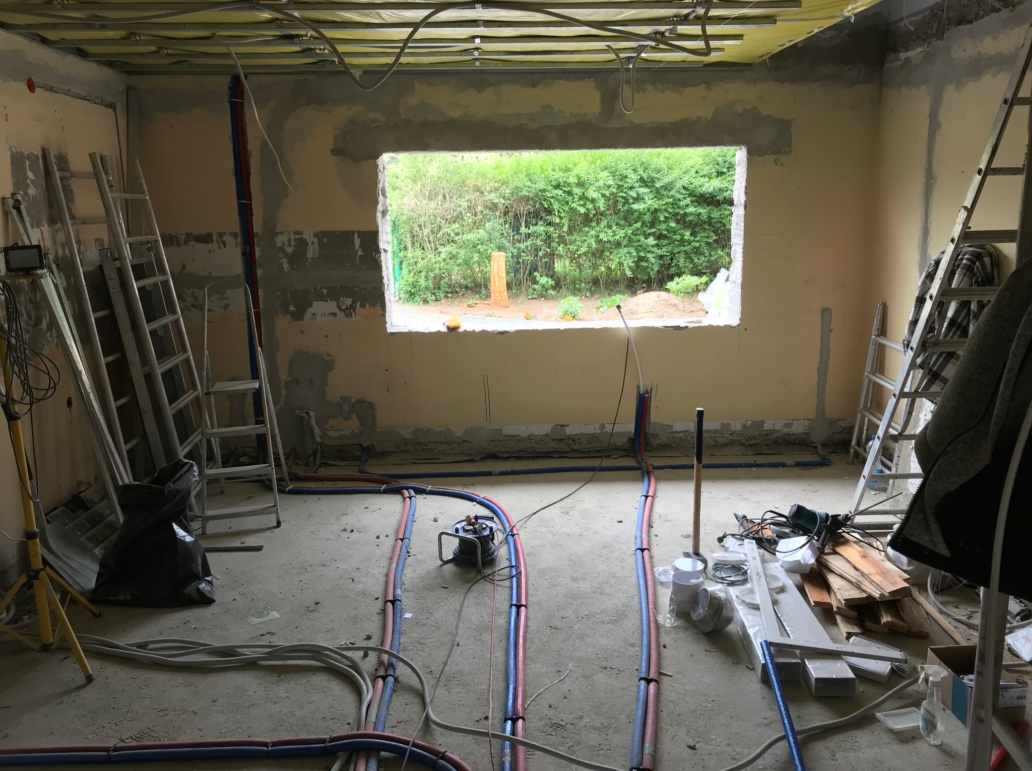 Wnętrze w trakcie remontu: widoczne instalacje, drabiny, narzędzia. Otwór okienny z widokiem na zieleń. Sufit z widoczną izolacją. Przewody instalacyjne na podłodze.