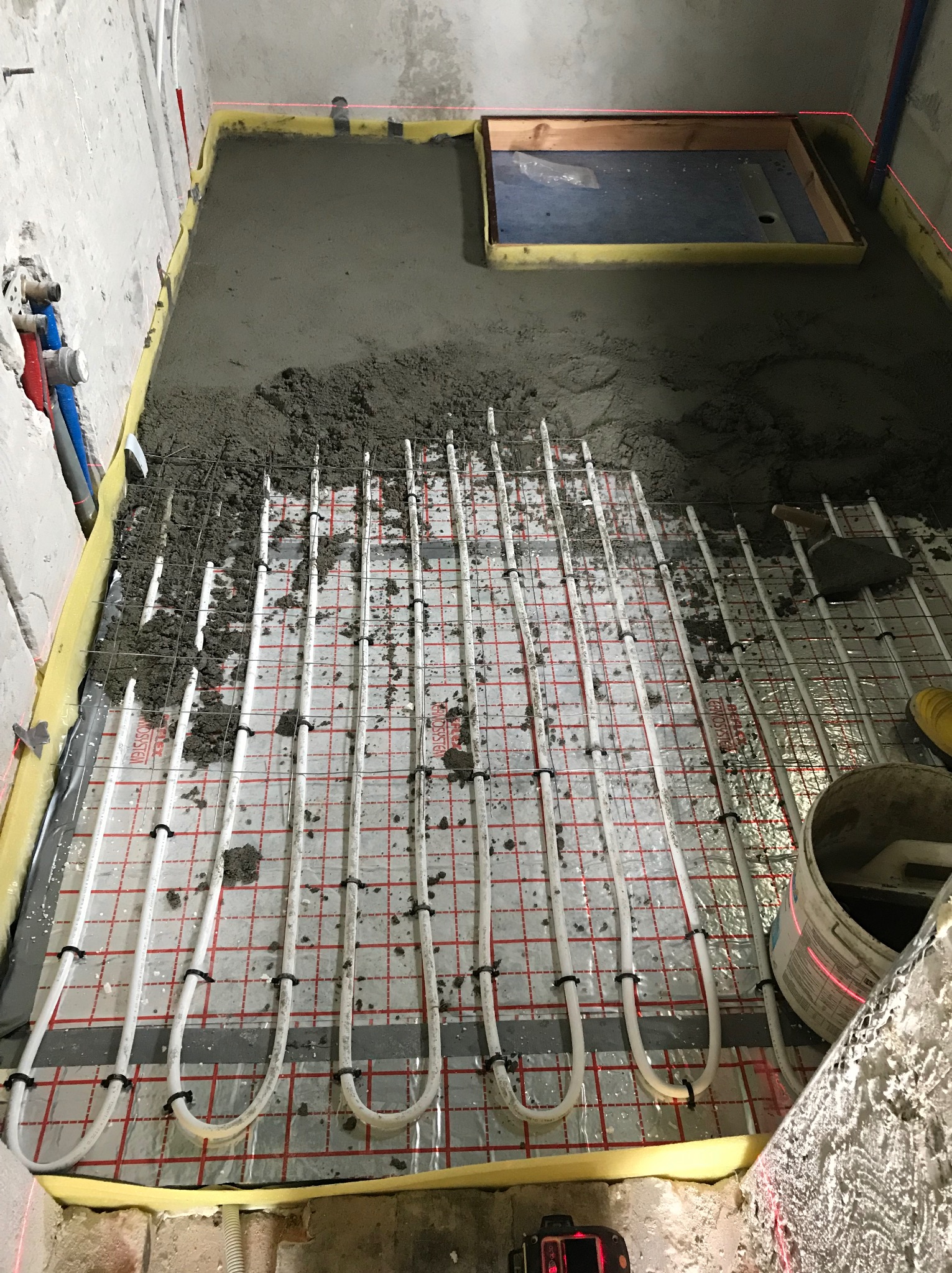 Montaż ogrzewania podłogowego w łazience: białe rurki na folii, częściowo zalane betonem, widoczny laser krzyżowy i wiadro z zaprawą. Etap prac przed położeniem płytek.