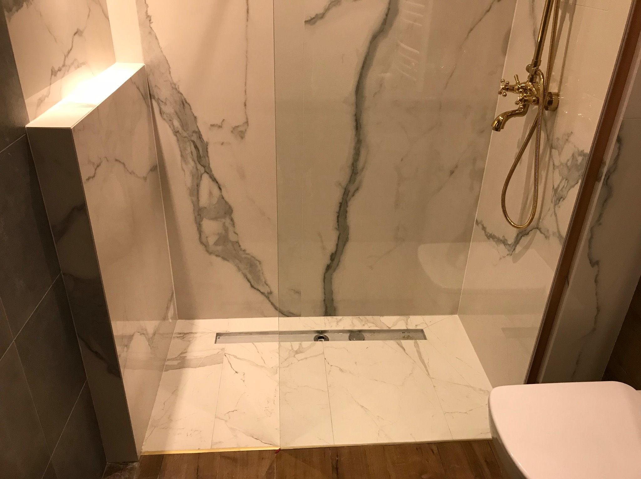 Wykończona kabina prysznicowa z białymi płytkami imitującymi marmur, złotą armaturą i odpływem liniowym. Widoczny fragment toalety.