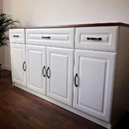 Biała komoda z ciemnobrązowym blatem, trzema szufladami i czterema drzwiczkami z czarnymi uchwytami, na tle drewnianej podłogi i rośliny doniczkowej.