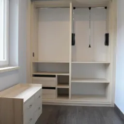 Jasna, drewniana szafa wnękowa z przesuwnymi drzwiami, półkami i szufladami, uzupełniona komodą w tym samym stylu, na tle ciemnej podłogi i jasnych ścian.