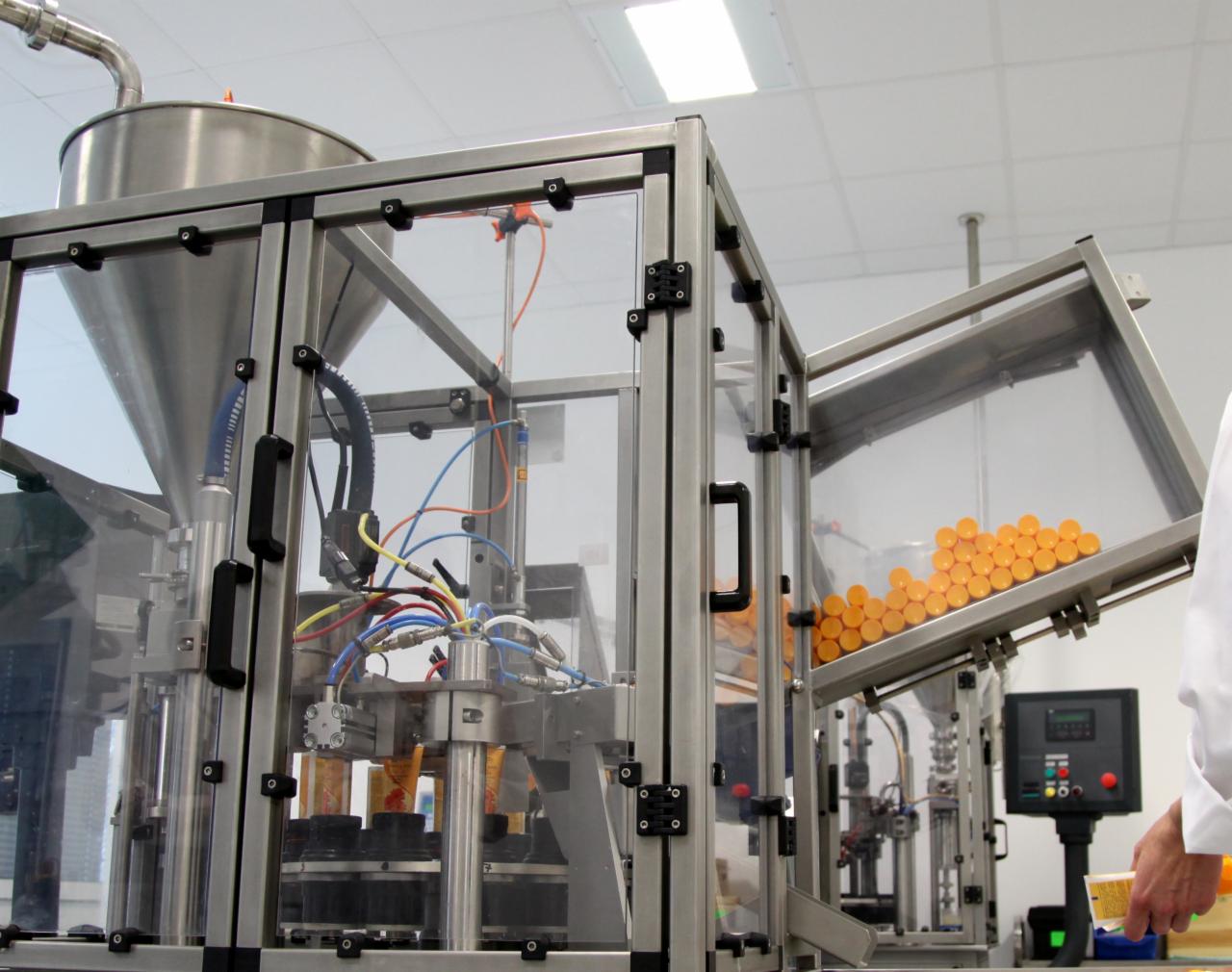 Automatyczna linia produkcyjna z przezroczystą obudową, dozownikiem, panelami sterowania i pomarańczowymi kapsułkami w trakcie pakowania; widoczny fragment ręki w białym fartuchu.