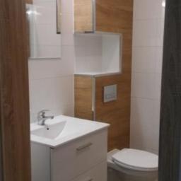 Usługi ogólno-budowlane - Nowoczesna łazienka po remoncie z białymi płytkami, drewnopodobnymi panelami na ścianie, wiszącą szafką z umywalką i toaletą