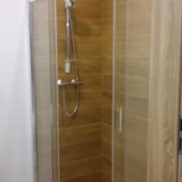 Usługi ogólno-budowlane - Kabina prysznicowa z przesuwnymi drzwiami, wyłożona brązowymi płytkami imitującymi drewno, z chromowaną armaturą i białą fugą, na tle białych płytek ściennych.