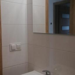 Usługi ogólno-budowlane - Biała umywalka z chromowaną baterią w nowoczesnej łazience z białymi, prostokątnymi płytkami i lustrem z drewnianą ramą, odbijającym drzwi z przeszkleniami.