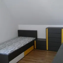 Pokój na poddaszu z łóżkiem, szafką nocną i komodą w kolorach szaro-żółto-białych, podłoga laminowana imitująca drewno, ściany białe.