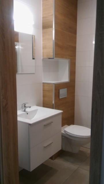 Nowoczesna łazienka po remoncie z białymi płytkami, drewnopodobnymi panelami na ścianie, wiszącą szafką z umywalką i toaletą