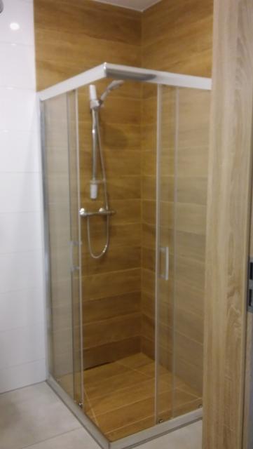 Kabina prysznicowa z przesuwnymi drzwiami, wyłożona brązowymi płytkami imitującymi drewno, z chromowaną armaturą i białą fugą, na tle białych płytek ściennych.