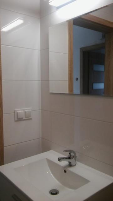 Biała umywalka z chromowaną baterią w nowoczesnej łazience z białymi, prostokątnymi płytkami i lustrem z drewnianą ramą, odbijającym drzwi z przeszkleniami.