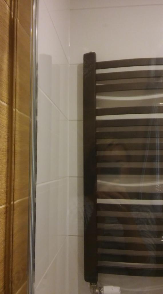 Fragment łazienki z brązowym grzejnikiem drabinkowym, białymi płytkami na ścianie i fragmentem drewnianej zabudowy prysznicowej.