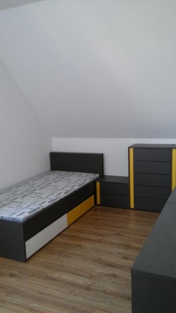Pokój na poddaszu z łóżkiem, szafką nocną i komodą w kolorach szaro-żółto-białych, podłoga laminowana imitująca drewno, ściany białe.