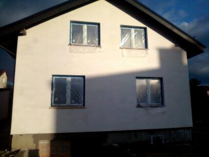 Dwukondygnacyjny dom jednorodzinny z nową, jasną elewacją, okna zabezpieczone folią malarską, widoczne prace wykończeniowe przy fundamentach.