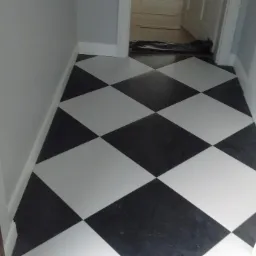 Czarno-biała podłoga w karo ułożona w wąskim korytarzu, widok z góry na świeżo położone płytki, z widocznym progiem i fragmentem otwartych drzwi.