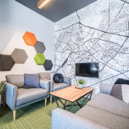 Salon z zieloną wykładziną dywanową w geometryczny wzór, szarą sofą, fotelami i dekoracyjną ścianą z mapą miasta oraz panelami akustycznymi w kształcie heksagonów.