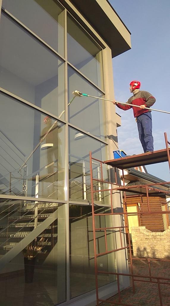 Pracownik w kasku na rusztowaniu myje długim mopem okna na wysokości w nowoczesnym budynku z przeszkloną klatką schodową.