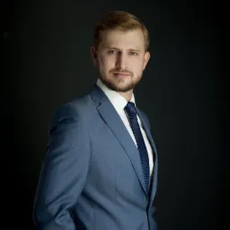 Portret młodego mężczyzny w błękitnym garniturze i granatowym krawacie na ciemnym tle, z delikatnym uśmiechem.