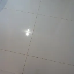 Błyszczące, białe płytki podłogowe ułożone w kwadratową siatkę, widoczne odbicie światła i fragment buta w rogu.