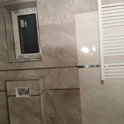 Wykończona łazienka z beżowymi płytkami imitującymi marmur, widoczne okno z białymi ramami, grzejnik drabinkowy i zabudowa stelaża WC z otwartym otworem rewizyjnym.