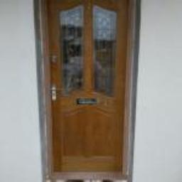 carpentry&joinery - Zewnętrzne, drewniane drzwi wejściowe z szybami o dekoracyjnym wzorze, zamontowane w jasnej ścianie. Widoczne elementy wykończeniowe wokół ościeżnicy i metalowa klamka.