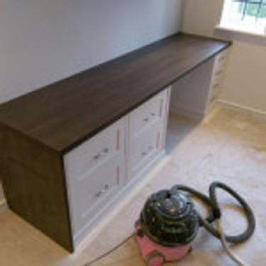 Długa, ciemna szafka z białymi frontami szuflad i blatem, stojąca pod oknem w jasnym pomieszczeniu, obok widoczny odkurzacz przemysłowy.