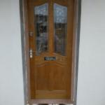 Zewnętrzne, drewniane drzwi wejściowe z szybami o dekoracyjnym wzorze, zamontowane w jasnej ścianie. Widoczne elementy wykończeniowe wokół ościeżnicy i metalowa klamka.