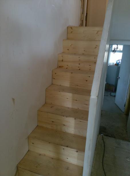 Surowe drewniane schody wewnętrzne w trakcie montażu, widoczne ślady ołówka i niedoskonałości obróbki drewna, biała ściana w tle z widocznymi niedoskonałościami tynku.