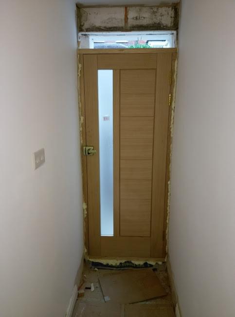 Wąskie przejście z nową, drewnianą ościeżnicą i drzwiami z długim, pionowym przeszkleniem, widoczne ślady pianki montażowej i niedokończone wykończenie ściany nad drzwiami.