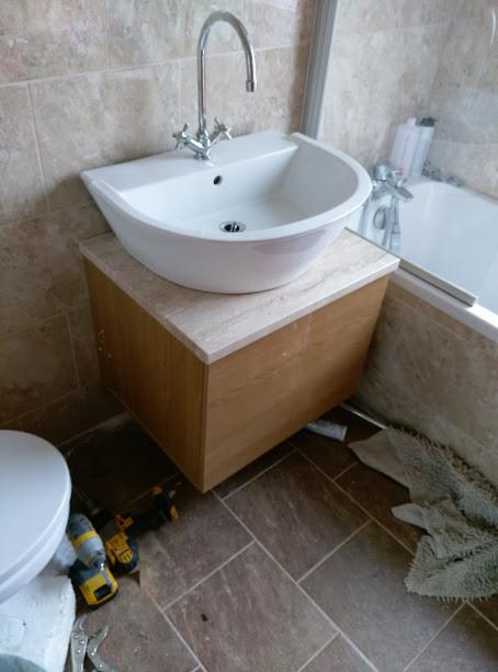 Umywalka nablatowa w kształcie łzy, zamontowana na szafce z drewnopodobnym wykończeniem, widoczna w łazience z beżowymi płytkami. Na podłodze leżą narzędzia i brudna ścierka.