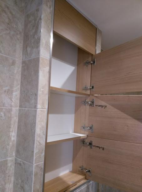 Otwarta szafka łazienkowa z jasnego drewna z białymi półkami, widoczne zawiasy i marmurowe płytki na ścianie po lewej stronie.