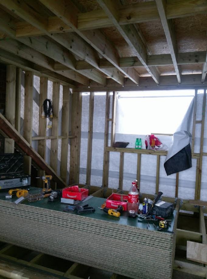 Surowy stan budowy: drewniana konstrukcja ścian i stropu z widocznymi narzędziami i materiałami budowlanymi wewnątrz pomieszczenia.