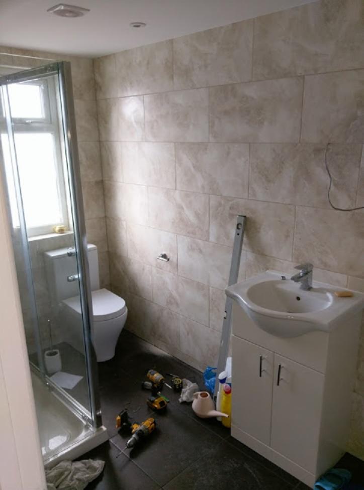 Ukończona łazienka z kabiną prysznicową, toaletą i umywalką z szafką, widoczne narzędzia i akcesoria na podłodze, płytki imitujące marmur na ścianach.