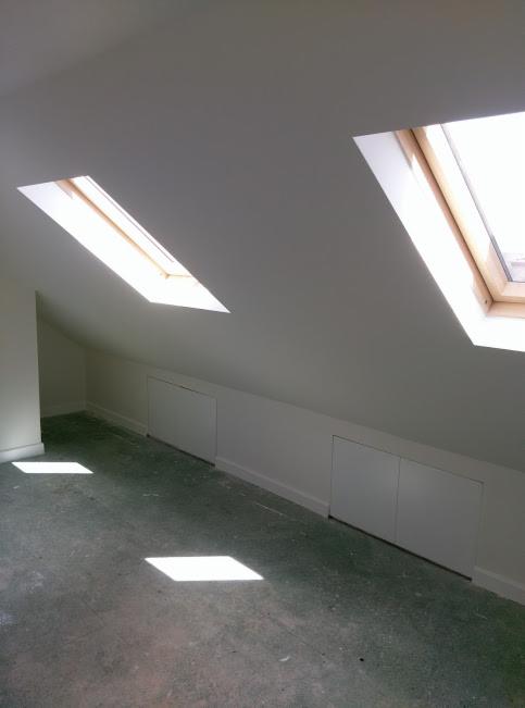 Poddasze z dwoma oknami dachowymi, widoczne białe ściany, skośny sufit i wbudowane szafki pod skosem, surowa podłoga w plamach światła słonecznego.