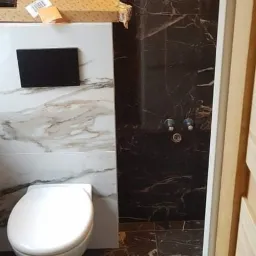 Wykończona łazienka z toaletą, czarnymi marmurowymi płytkami na ścianach i podłodze, oraz biało-marmurową zabudową toalety z czarnym przyciskiem spłukiwania. Na zabudowie toalety widoczny karton...