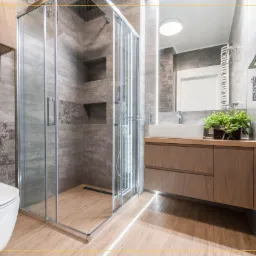 Nowoczesna łazienka z kabiną prysznicową typu walk-in, umywalką nablatową z drewnianą szafką, szarymi płytkami z dekoracyjnym pasem i drewnopodobną podłogą.