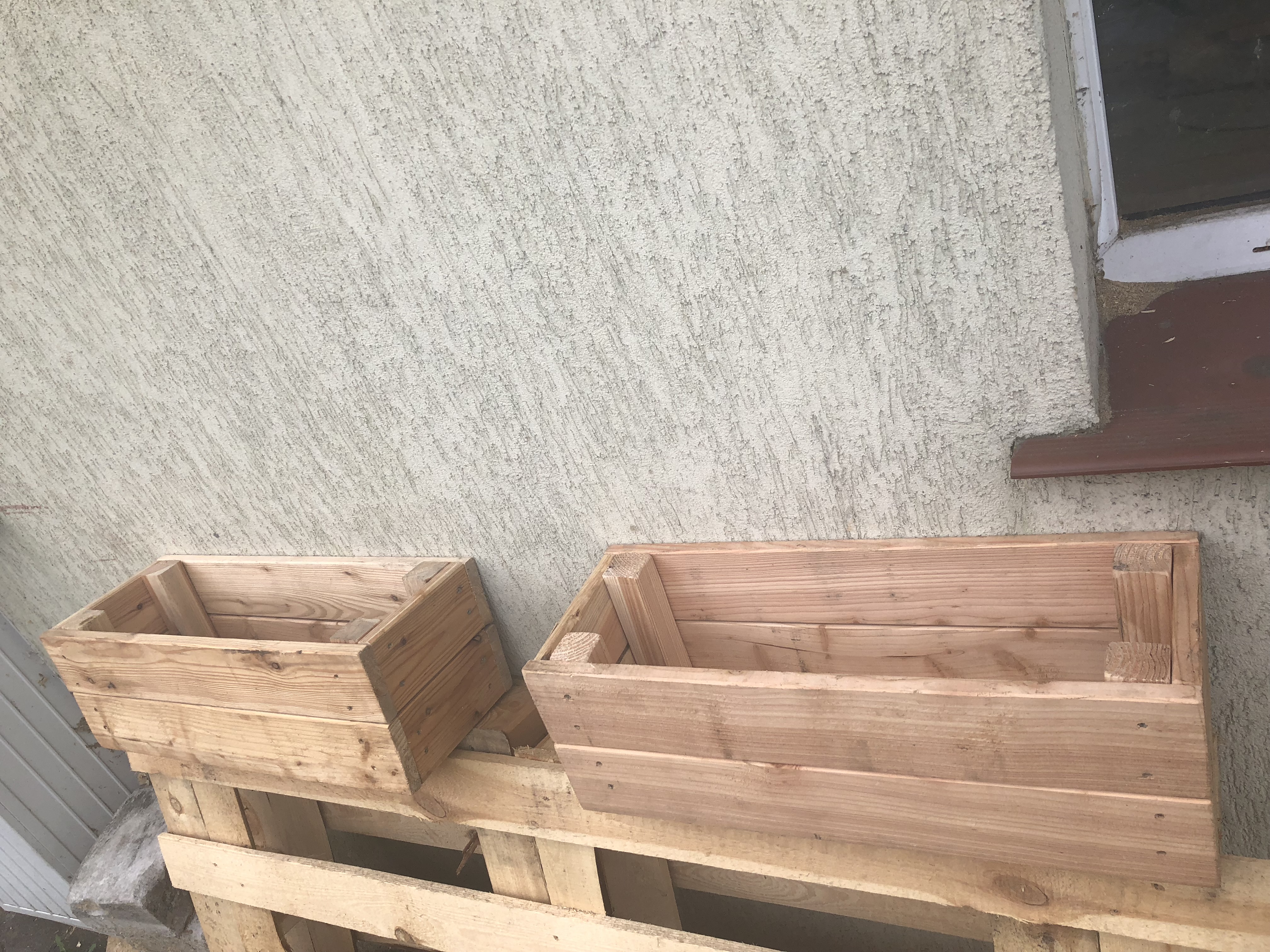 Dwie surowe, drewniane skrzynki na kwiaty ustawione na palecie przed szarą ścianą z widocznym parapetem okna.