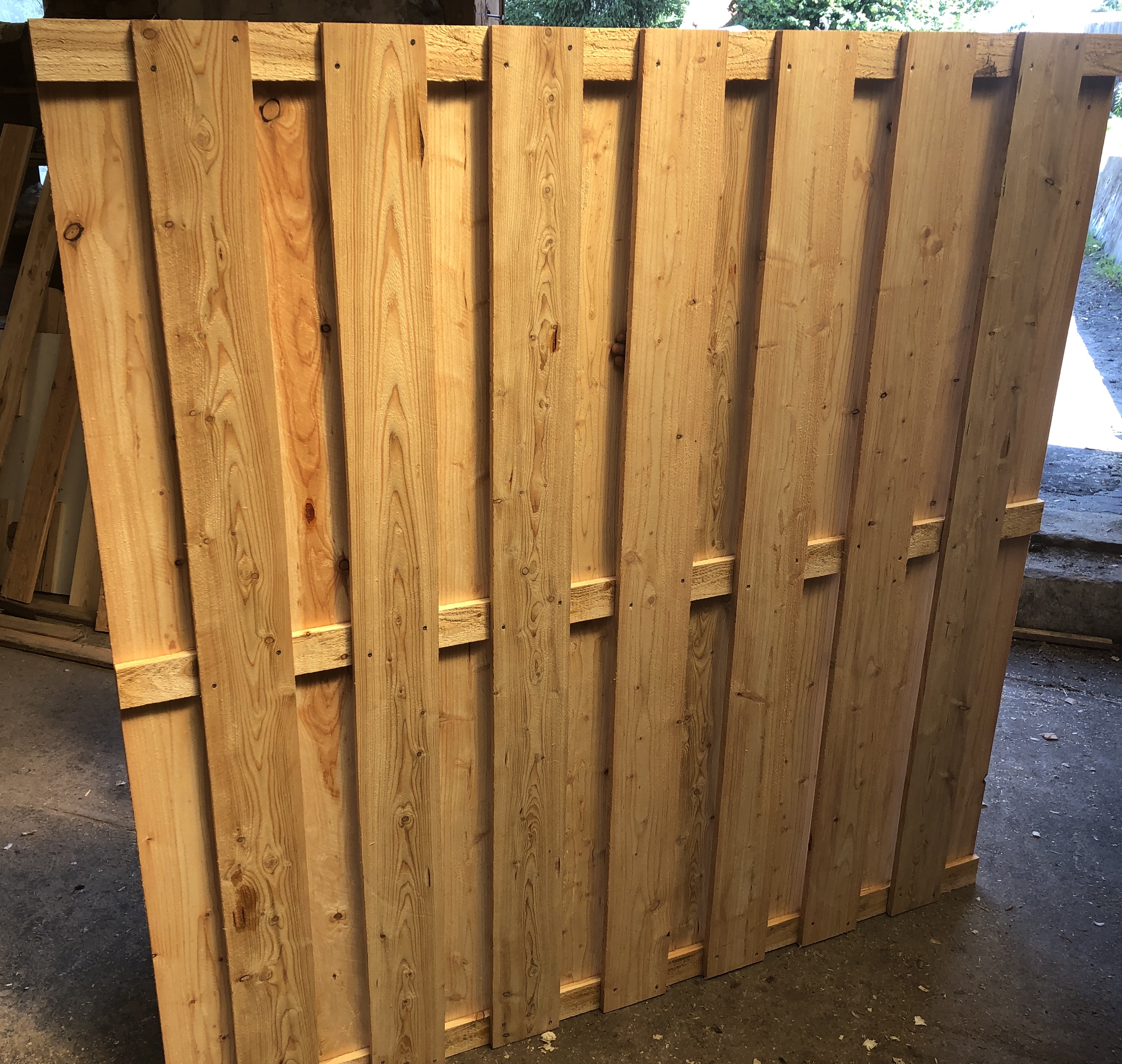 Surowe, jasne deski na płot, połączone poprzecznymi listwami, z widocznymi śladami obróbki i gwoździami, w tle składowane inne elementy drewniane.