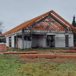 Surowy stan budowy parterowego domu z bloczków, z widoczną konstrukcją dachu z drewnianych belek, materiały budowlane składowane obok